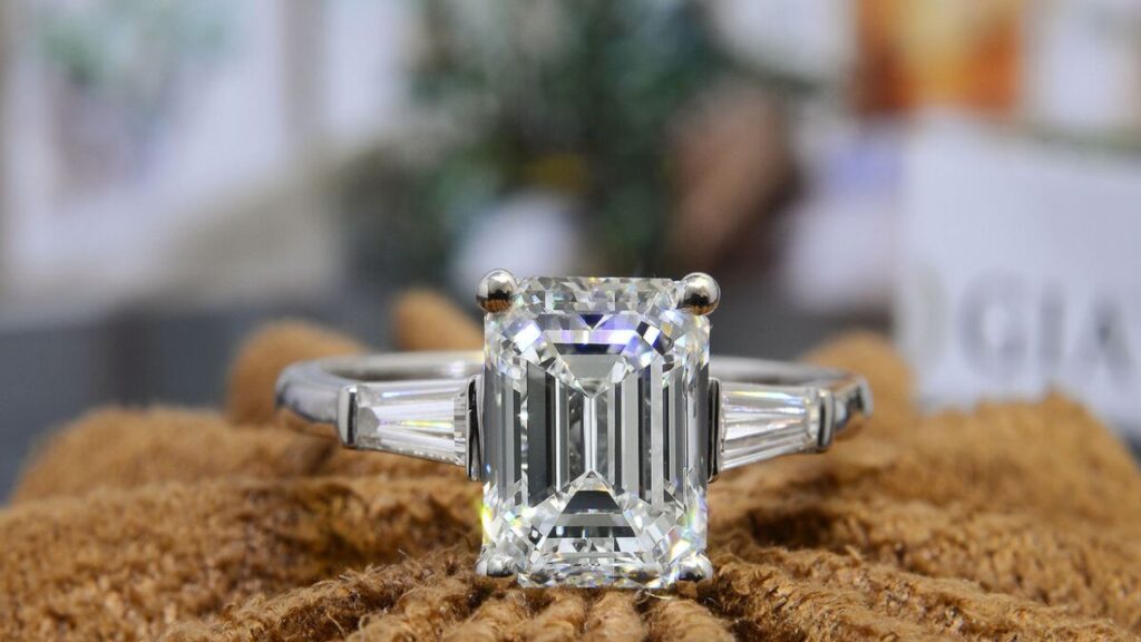 A platinum ring featuring a large emerald-cut diamond flanked by two baguette diamonds, displayed on a textured, neutral-colored surface.