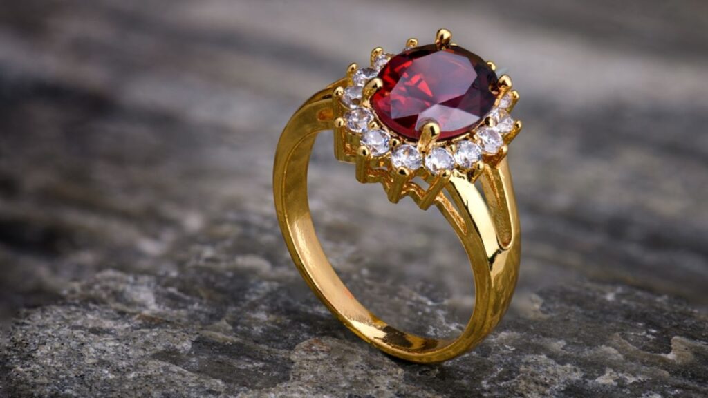 A gold ring with a large, oval-shaped red gemstone surrounded by a halo of smaller white gemstones, resting on a textured gray surface.