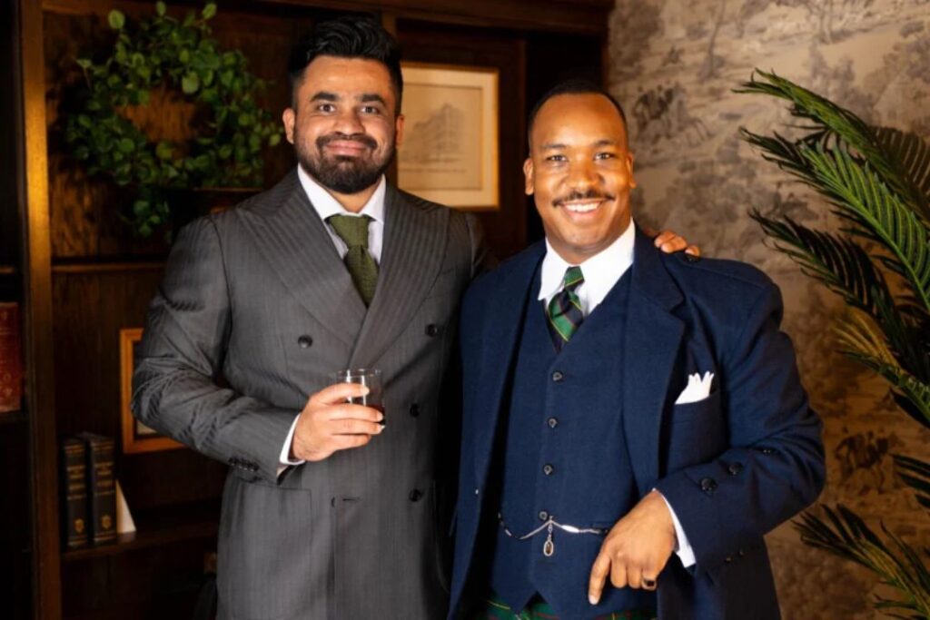 Two sharply dressed men in tailored suits smile confidently in a warm, elegant room with wood-paneled walls, books, and lush greenery.
