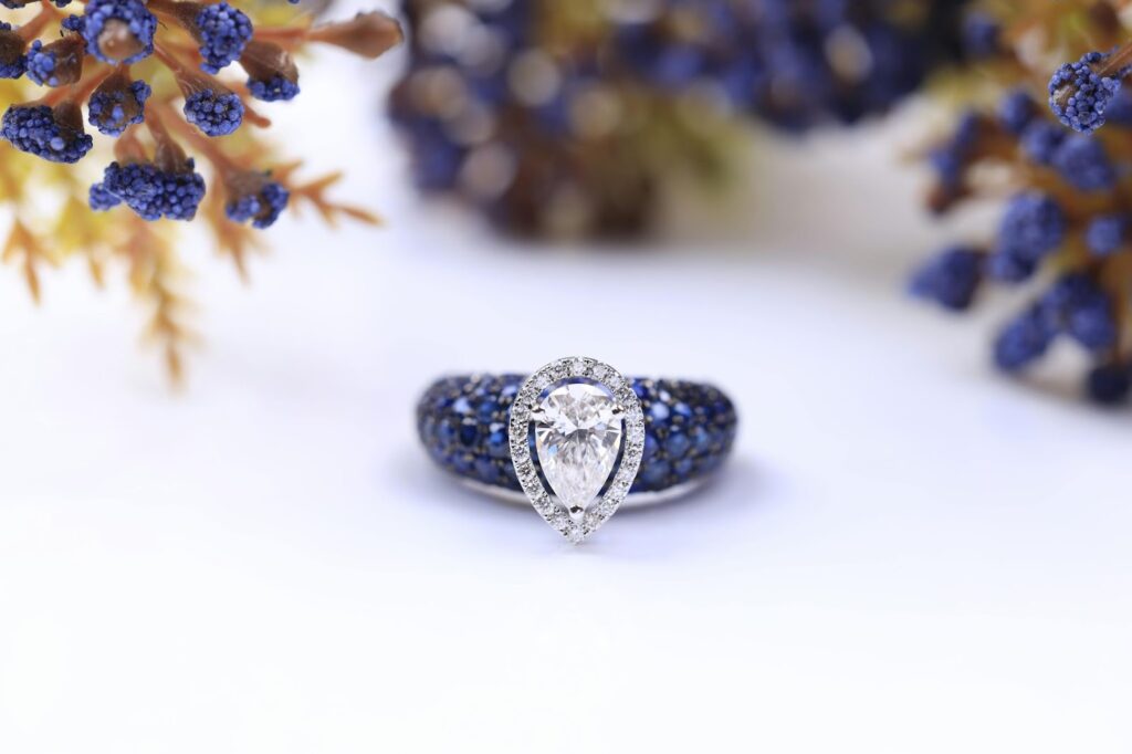 A unique diamond ring featuring a pear cut center stone surrounded by a diamond halo, and sapphires set in the band.