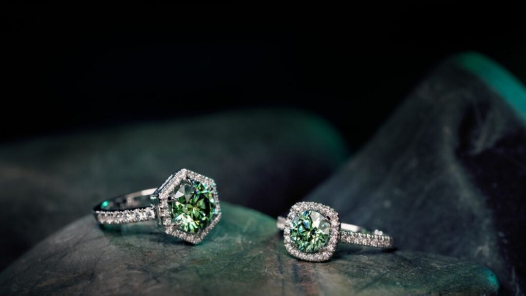 Two green diamond rings, one with a hexagonal setting and the other with an oval setting, both with diamond halos, resting on a dark, textured surface.