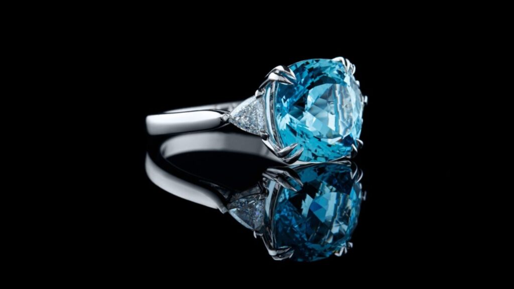 A platinum ring featuring a large, cushion-cut blue topaz gemstone flanked by two smaller triangular diamonds, reflected on a black surface.