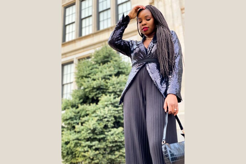 A stylish woman in a sequined blazer and pleated pants poses confidently outside a classic building, holding a black handbag with braided hair.