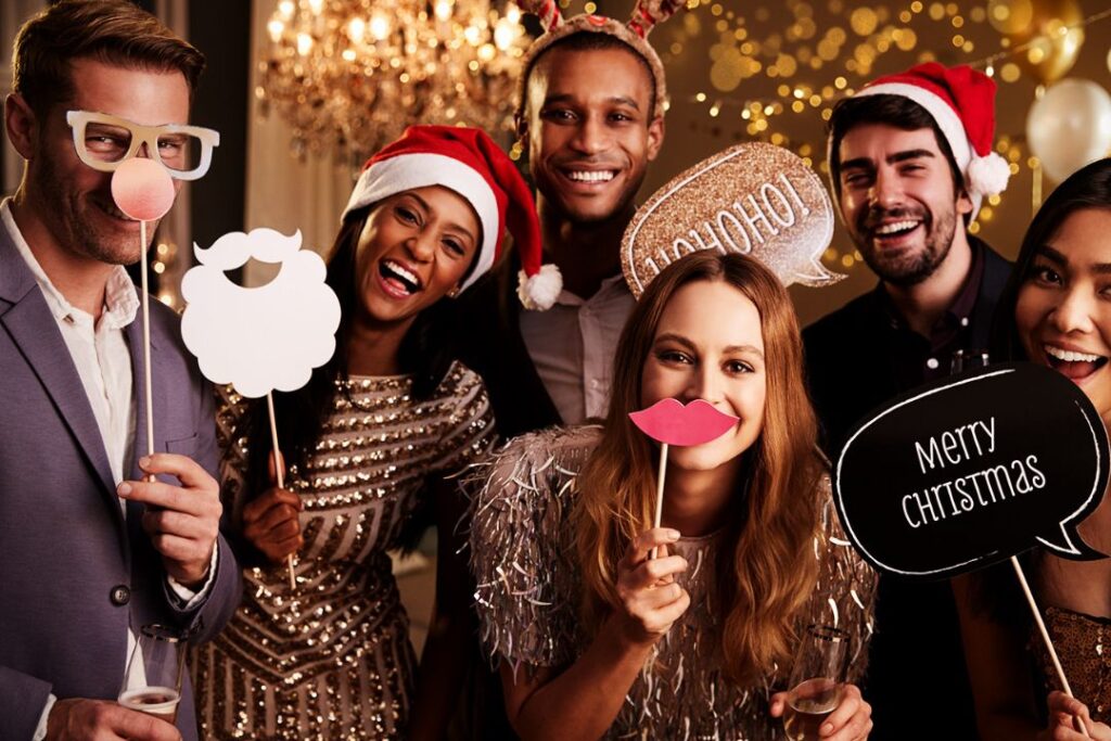 A joyful group at a festive Christmas party, dressed in holiday attire, holding playful props with smiles and laughter under twinkling lights.