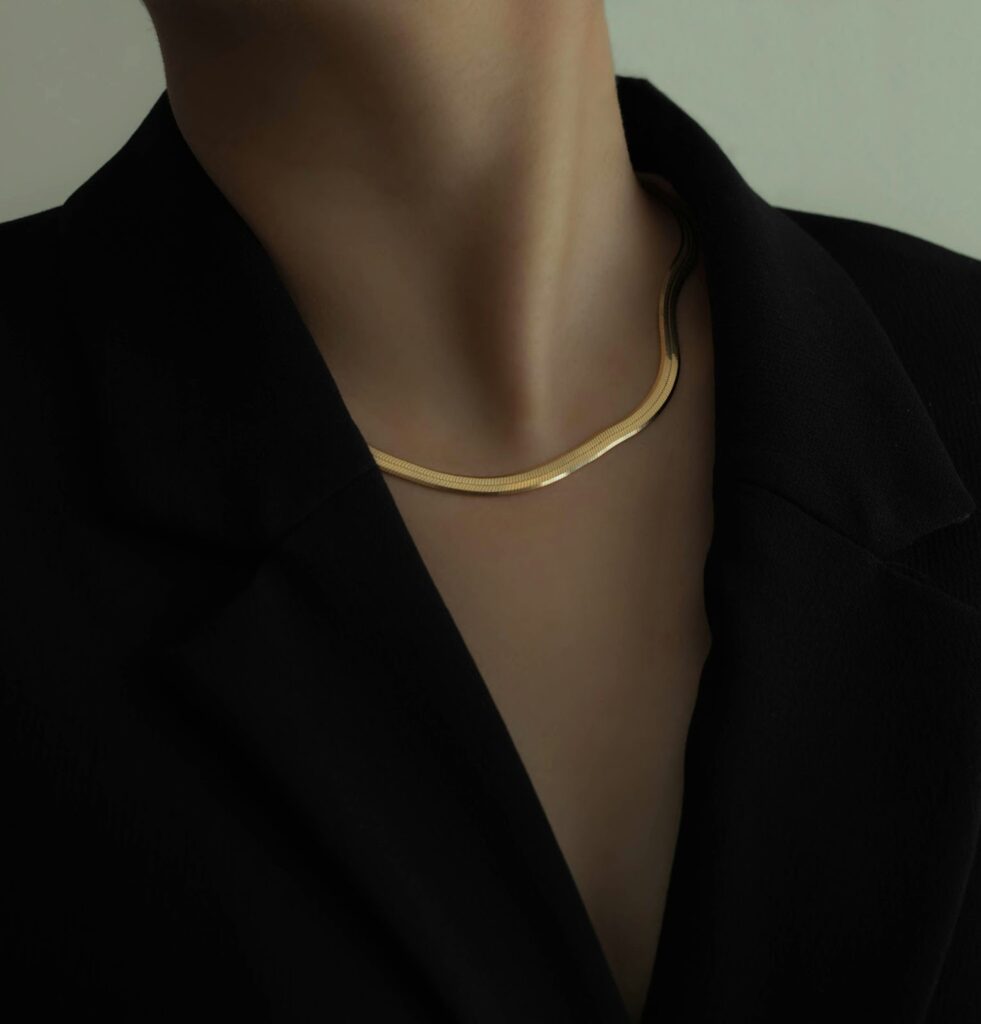 A close-up of a gold herringbone chain necklace on a person wearing a black jacket, focusing on the necklace's texture and shine.