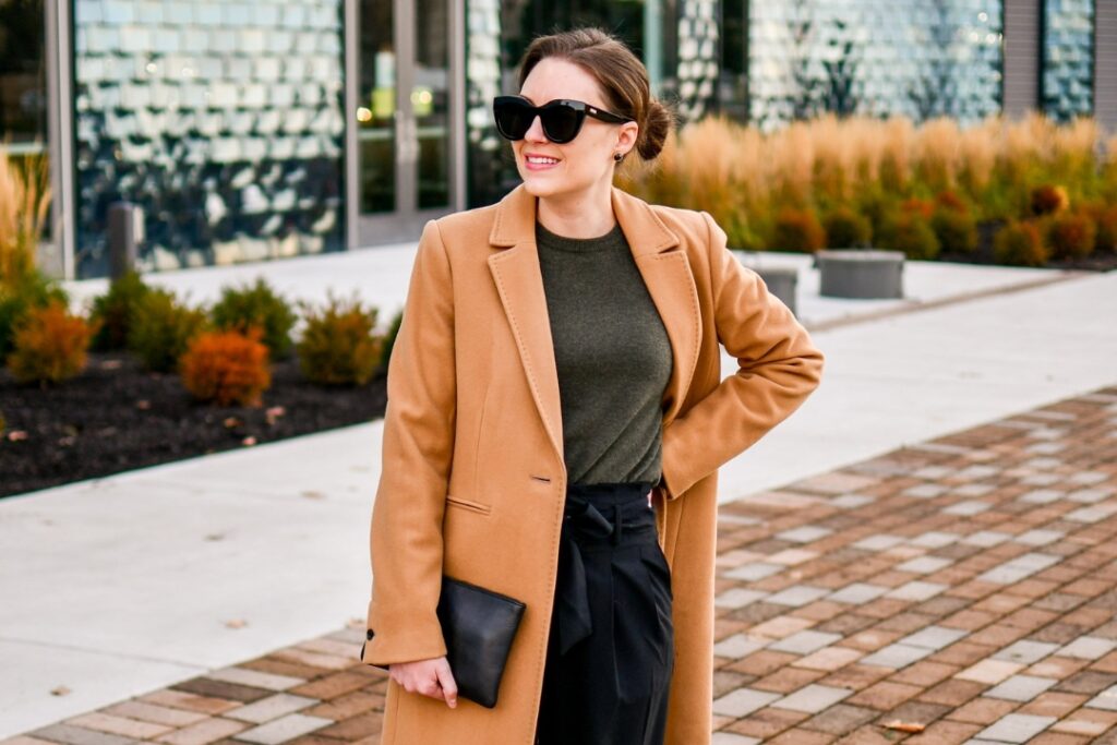 A stylish woman in a camel coat and sunglasses walks confidently on a brick pathway, with a modern glass building reflecting the sky.