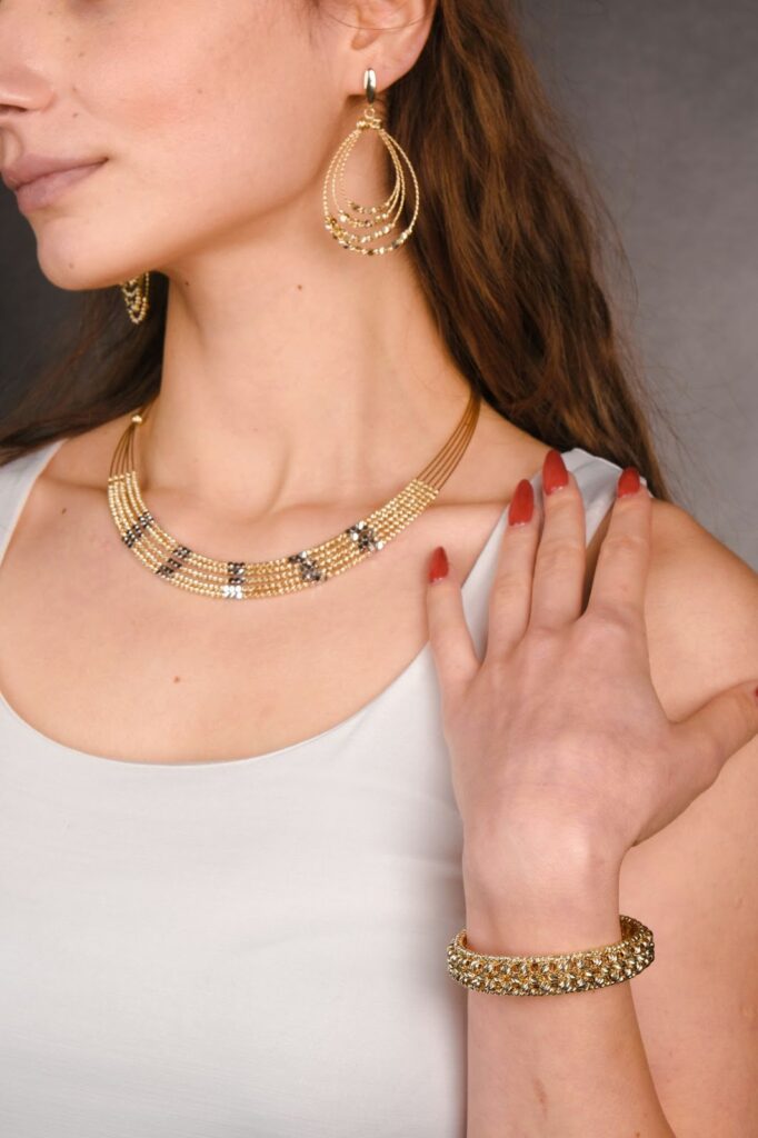 A woman wearing gold jewelry, including a necklace, earrings, and a bracelet, poses against a dark gray background.