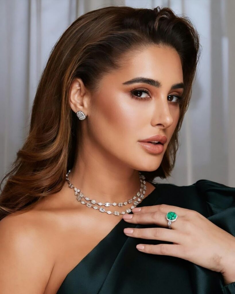 A woman with long brown hair, wearing a green dress and diamond jewelry, poses with a serious expression against a light background.