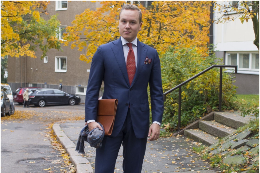 A man in a navy blue suit and red tie stands outdoors, holding a brown folder and gray scarf, with autumn foliage in the background.
