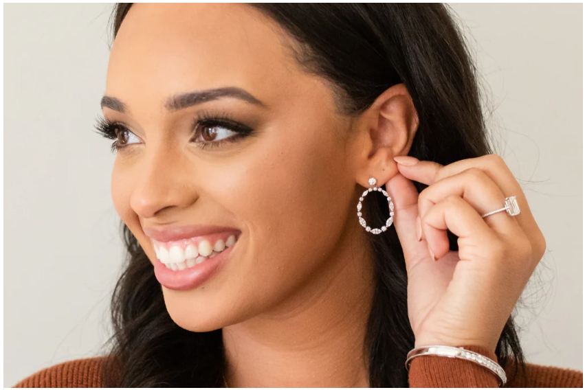A woman with dark hair and a warm smile showcases oval diamond earrings, a diamond ring, and a silver bracelet against a light background.