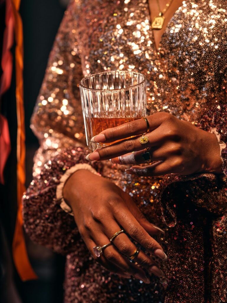 A person in a sequined dress holds a drink, adorned with various rings and a necklace. Not typically Professional Jewelry For Work.