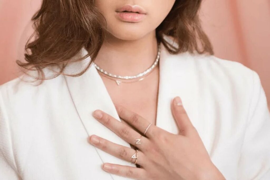 A person wearing delicate necklaces and rings with letters, showcasing Office Jewelry Essentials.