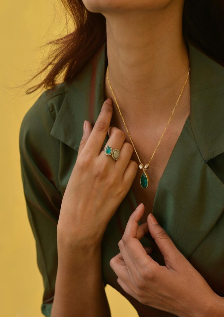 A person wearing a gold necklace with a green pendant and a ring with green stones, Professional Jewelry For Work.