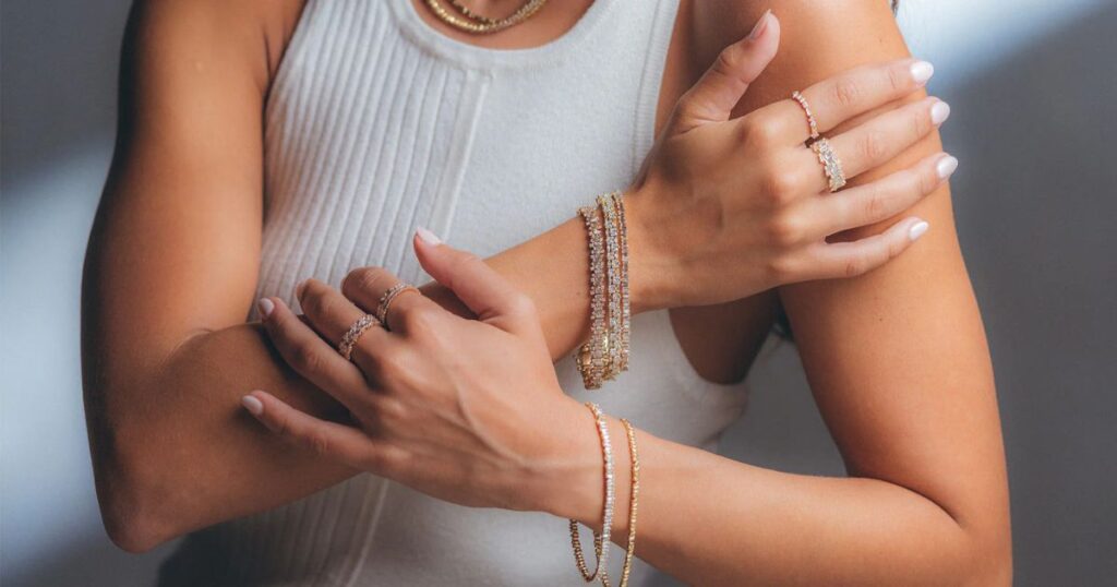 A person's crossed arms with multiple bracelets and rings are shown against a beach background, showcasing Stylish jewelry for holidays from Wulf Diamond Jewelers.