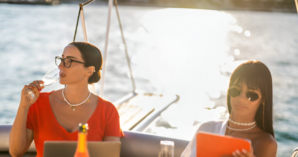 A woman on a yacht sips champagne, wearing a delicate necklace from Wulf Diamond Jewelers' Cruise-friendly jewelry collection.