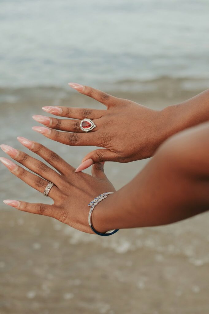 A person's hands with rings and a bracelet are posed over a blurred beach background, showcasing Summer vacation accessories from Wulf Diamond Jewelers.