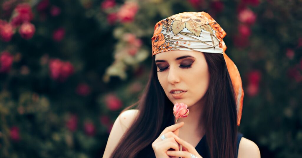 A woman with long hair, a patterned headscarf, and rings holds a flower, embodying the spirit of Summer vacation accessories from Wulf Diamond Jewelers.
