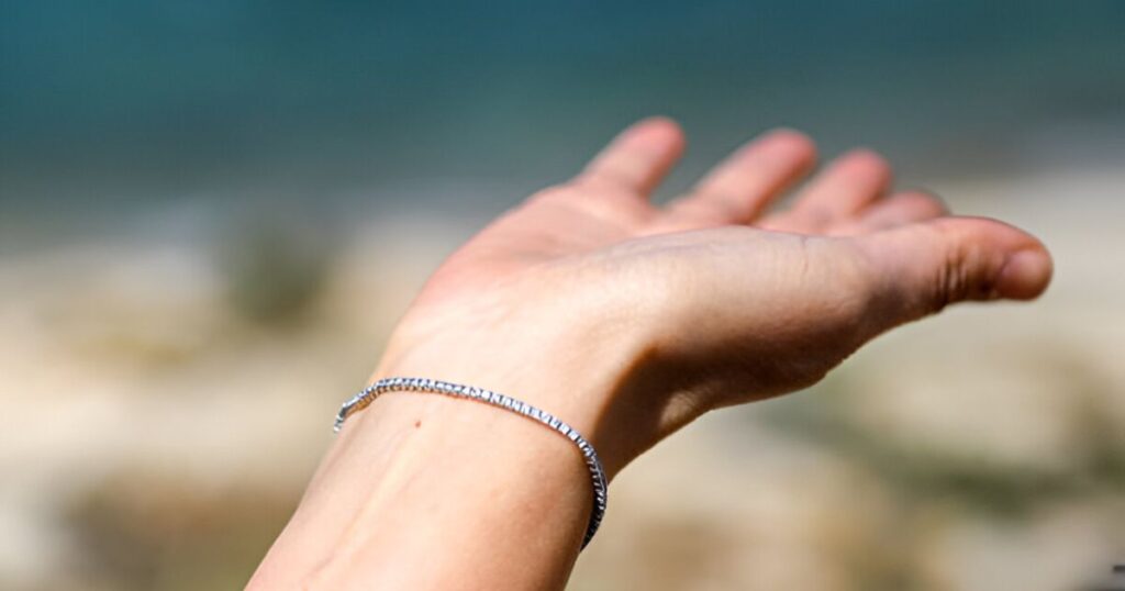 A hand reaches towards blurred beach scenery, wrist adorned with a delicate bracelet, representing Wedding jewelry for destination events from Wulf Diamond Jewelers.