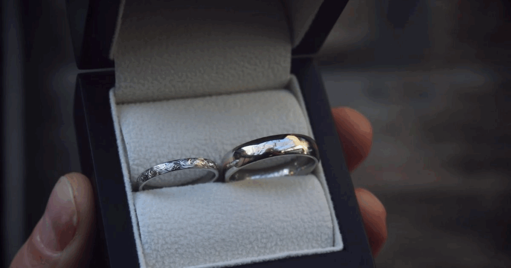 
A hand holds an open black jewelry box showing two silver wedding rings, featuring hypoallergenic jewelry for travel from Wulf Diamond Jewelers.